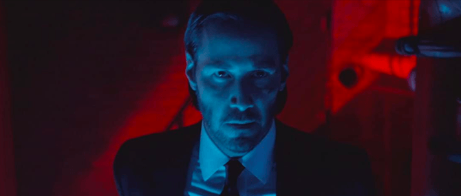 A movie still from John Wick shows John Wick (played by Keanu Reeves) bathed in a blue light. Behind him is a red light pouring out of a window.