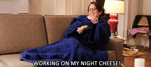 A GIF from the TV show 30 Rock where Tina Fey’s character Liz Lemon wears a blue slanket and eats cheese she just peeled off of the block. She’s singing “Working on my night cheese!”