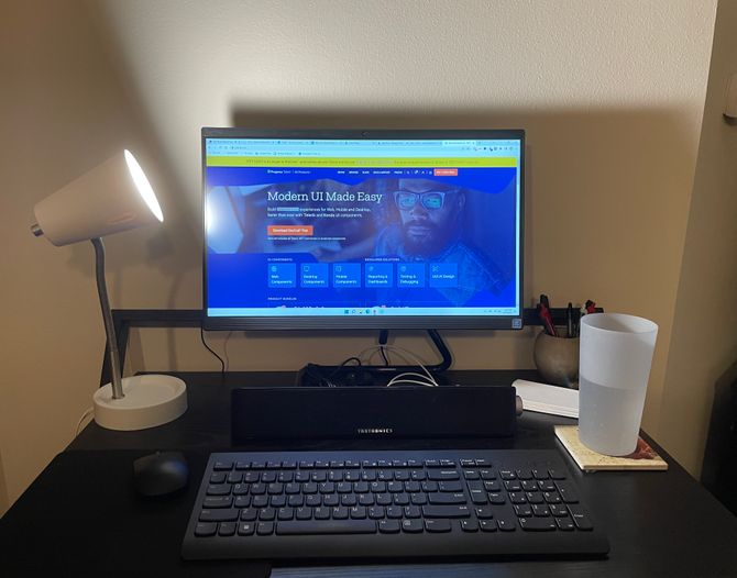 A black desk with a simple workplace setup. A small white lamp sits on the left side of the desk along with a mouse and mousepad. In the center is an all-in-one desktop computer, a Taotronics speaker, and a black keyboard. On the right is a jar of pens, some stationery, and a cup of water on a coaster.