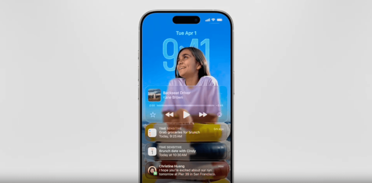 In this Liquid Glass iPhone lock screen example, we see a smiling girl in front of the time (which is Liquid Glass). In front of the girl is an audio player that says “Backseat Driver” by Kane Brown along with 3 notifications for a tasks, calendar appointment, and text. These 3 cards and audio player are semi-transparent.