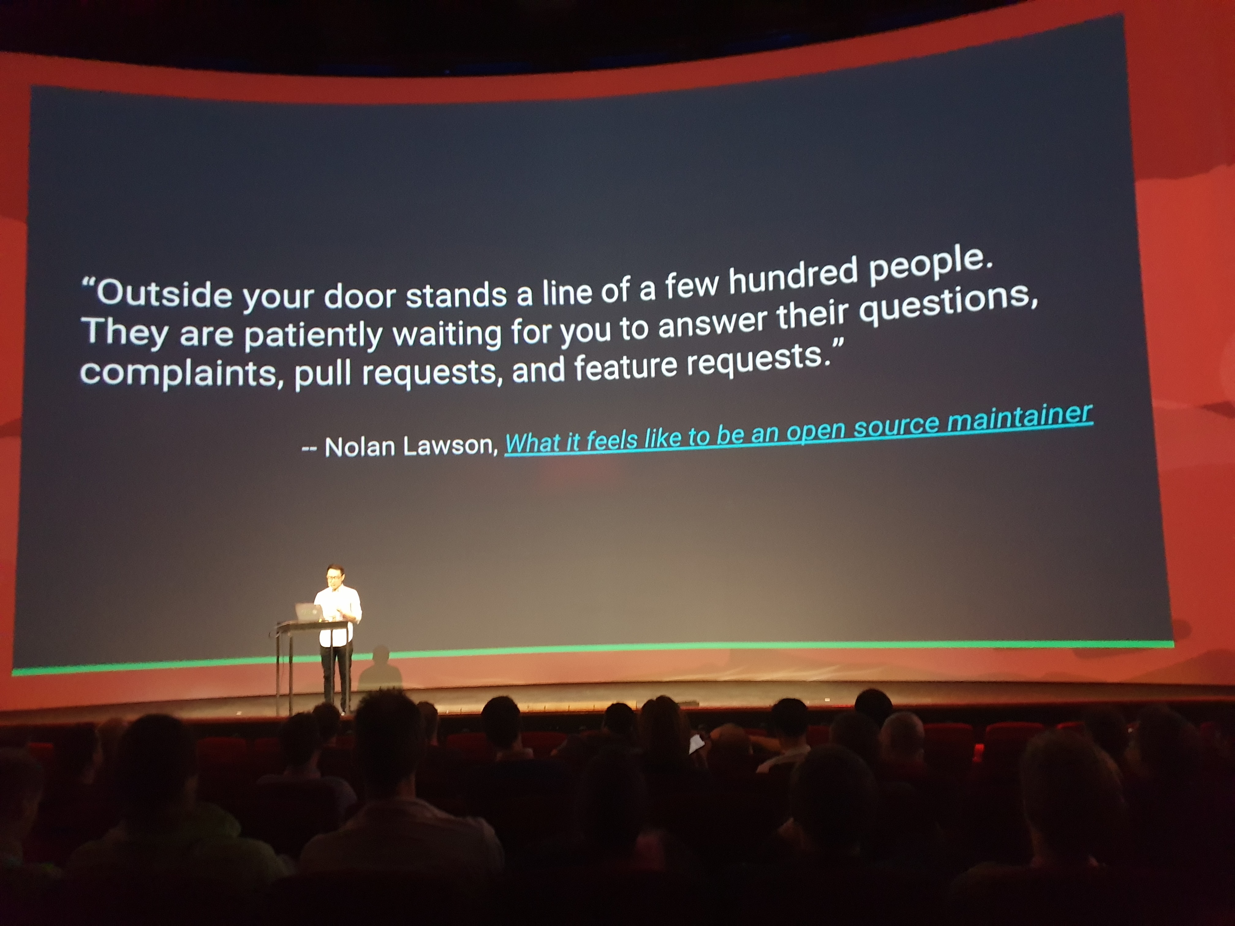 Evan You on stage in front of a slide with a quote by Nolan Lawson. It reads, “Outside your door stands a line of a few hundred people. They are patiently waiting for you to answer their questions, complaints, pull requests, and feature requests.”